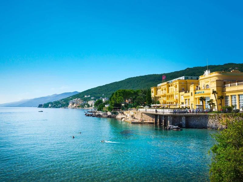 Wybrzeże z czystą niebieską wodą, budynki nad brzegiem i zalesione wzgórza w tle.