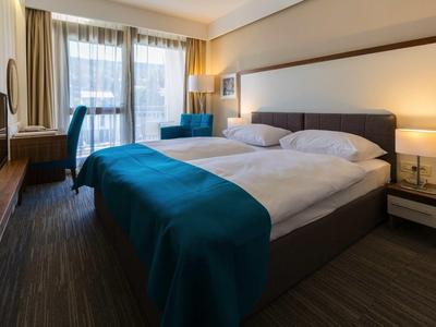 Nowoczesny pokój hotelowy z dużym łóżkiem podwójnym, biurkiem i częścią wypoczynkową przy naturalnym świetle.