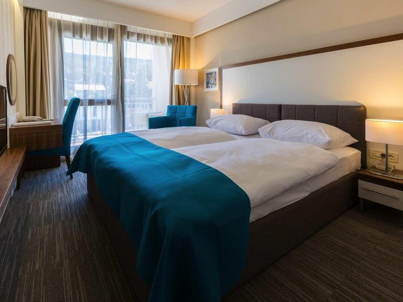 Nowoczesny pokój hotelowy z dużym łóżkiem podwójnym, biurkiem i częścią wypoczynkową przy naturalnym świetle.