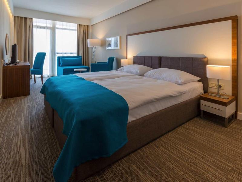 Nowoczesny pokój hotelowy z dużym łóżkiem, niebieskimi akcentami i miejscem do siedzenia przy oknie.