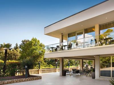 Edificio de hotel moderno con grandes ventanas de vidrio y balcón en entorno soleado con árboles.