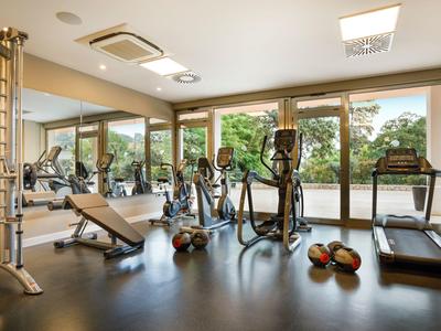 Moderner Fitnessraum mit Geräten und großen Fenstern, die auf grüne Bäume blicken.