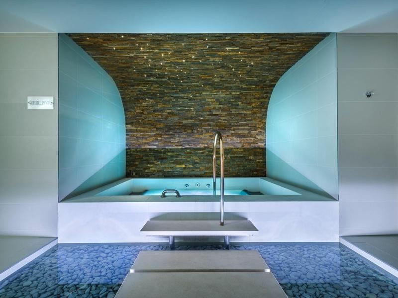 Piscina de hotel moderna con suelo de mosaico y pared trasera de piedra redondeada.