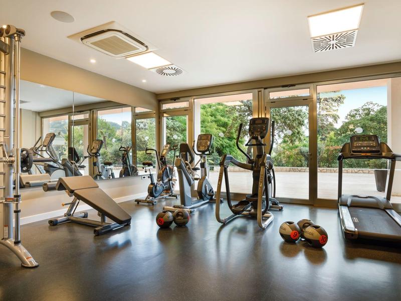 Gimnasio con equipos modernos y grandes ventanas con vista a árboles verdes.