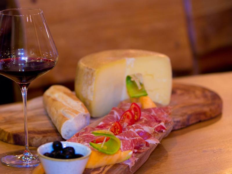 Tabla de madera con embutidos y queso, aceitunas y una copa de vino tinto sobre una mesa.