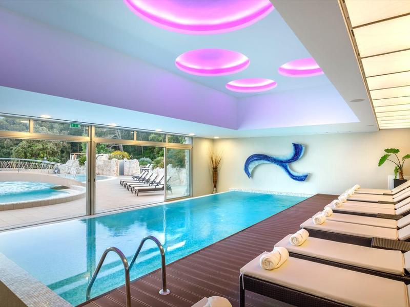 Piscina interior moderna con tumbonas, ventanas panorámicas y vista a la piscina exterior y montañas.