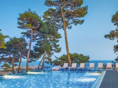 Zwembad met ligstoelen met uitzicht op zee, omringd door dennenbomen onder een blauwe lucht.