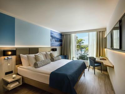 Moderní hotelový pokoj s manželskou postelí, židlí, psacím stolem a výhledem na moře přes velká okna.