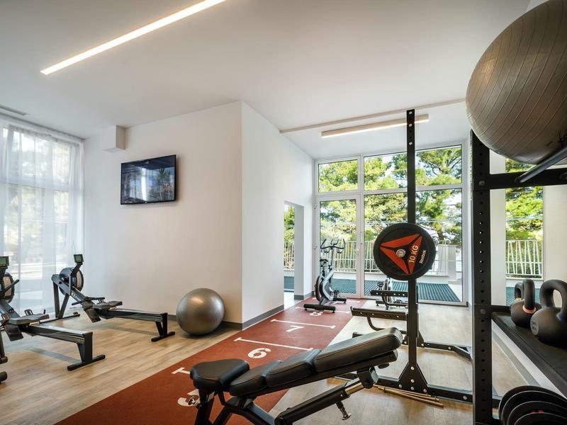 Moderní posilovna s kardio zařízením, činkami a lavicí v světlé místnosti.