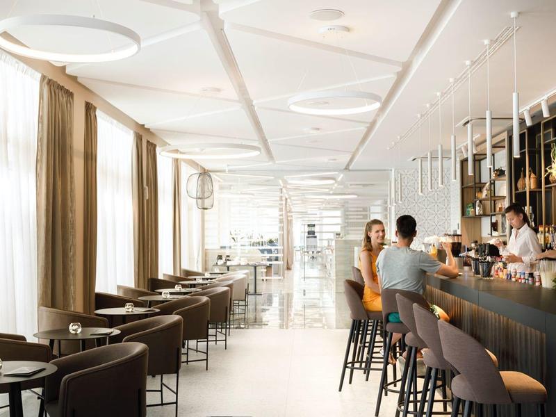 Moderní hotelová restaurace s barem, posezením a velkými okny pro denní světlo.