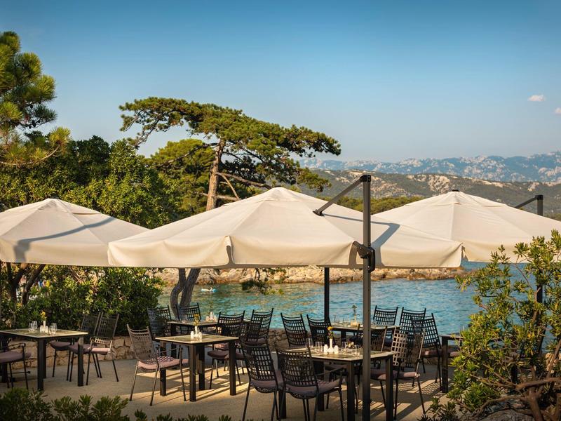 Venkovní restaurace s bílými slunečníky a stoly u jezera a hor.