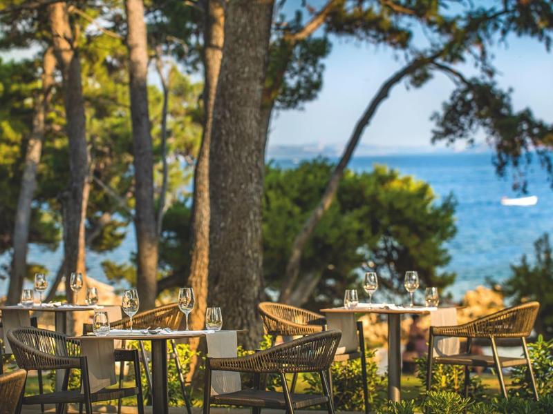 Venkovní stoly a židle pod stromy s výhledem na moře a plachetnici.