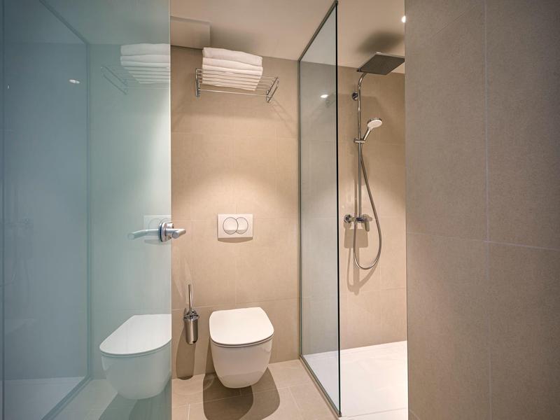 Moderní hotelová koupelna s bezbariérovým sprchovým koutem, toaletou a béžovými dlaždicemi.