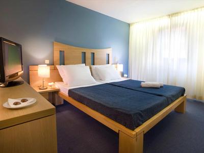 Eenvoudig ingericht hotelkamer met tweepersoonsbed, tv en grote ramen met gordijnen.