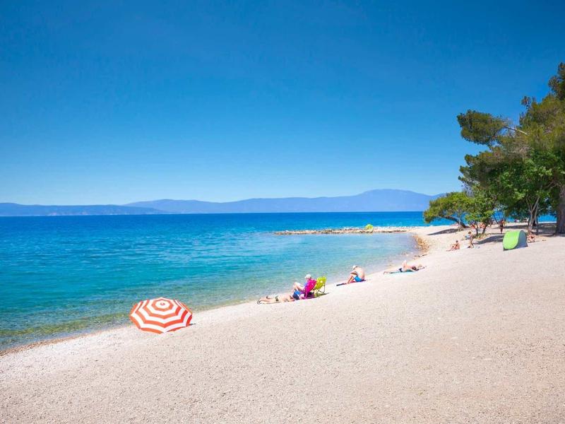 Sunny beach with clear blue sea, white pebbles, beach umbrellas, and trees in the background.