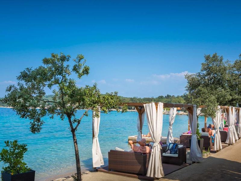 Seaside lounge area with cushioned seats and white drapes under clear blue sky.