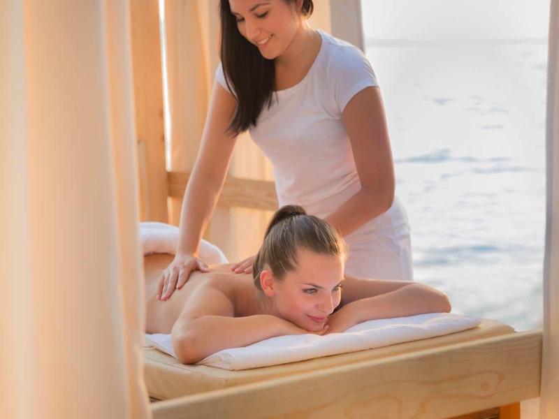 A woman receiving a back massage in a seaside spa room with soft curtains and natural light.