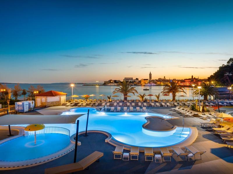 Wieczorny widok na oświetlony basen i leżaki z morzem i miastem w tle.