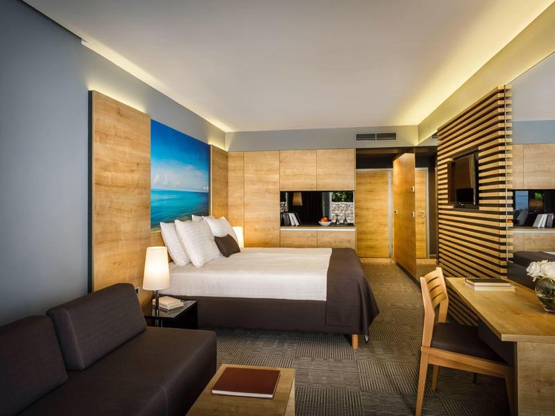 Nowoczesny pokój hotelowy z łóżkiem podwójnym, sofą i biurkiem w ciepłych drewnianych odcieniach.