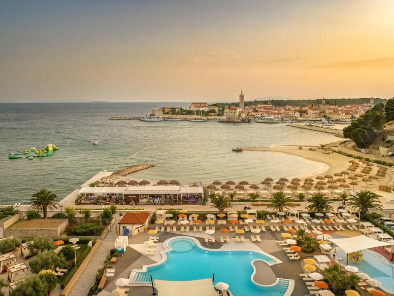 Zachód słońca nad hotelem przy plaży z basenem, leżakami i widokiem na nadmorskie miasto.