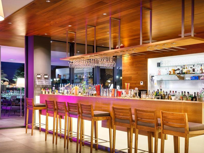 Nowoczesny bar hotelowy z drewnianymi detalami i wysokimi stołkami, oświetlony ciepłym światłem.