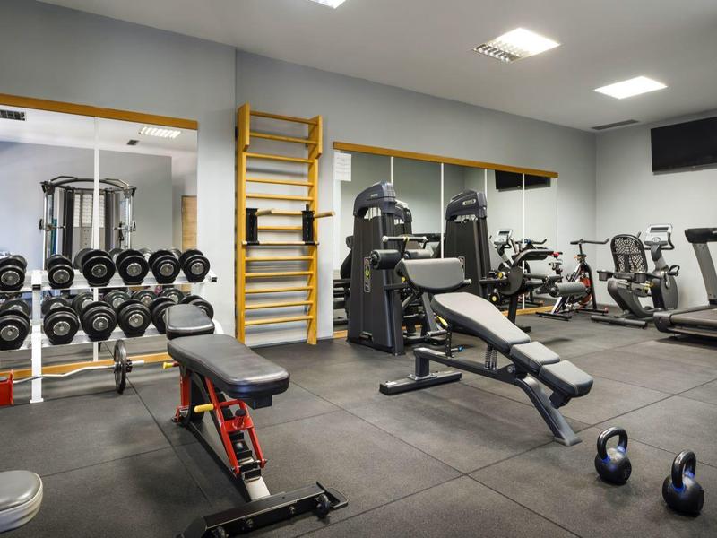 Nowoczesna siłownia z hantlami, ławkami i sprzętem przed dużym lustrem.