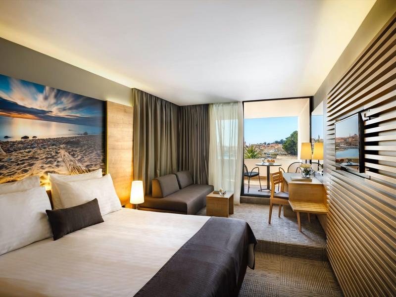 Nowoczesny pokój hotelowy z podwójnym łóżkiem, dużym obrazem na ścianie i balkonem z widokiem na miasto.