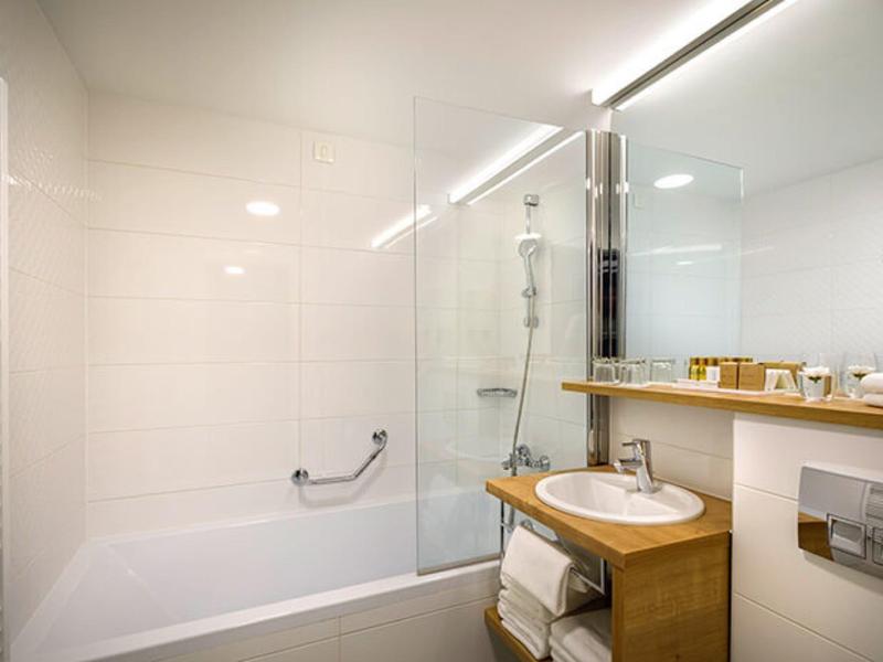 Jasna łazienka z wanną, szklaną kabiną prysznicową i drewnianą szafką pod umywalką.