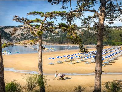 Zandstrand met parasols bij het meer, omgeven door bomen en heuvels op de achtergrond.