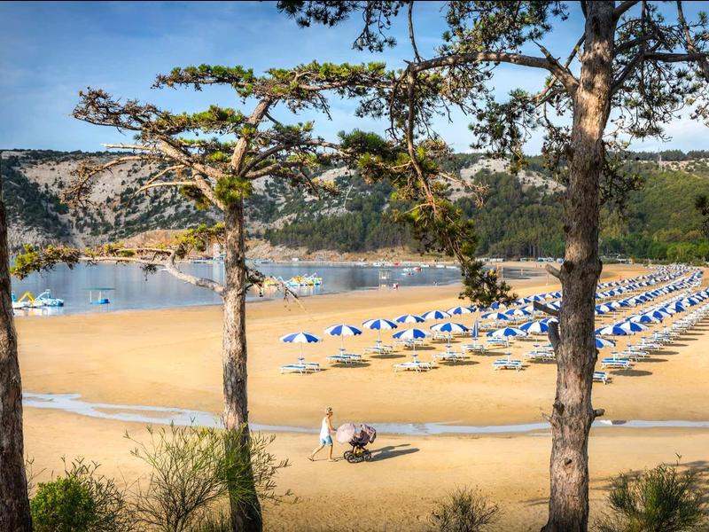 Zandstrand met parasols bij het meer, omgeven door bomen en heuvels op de achtergrond.