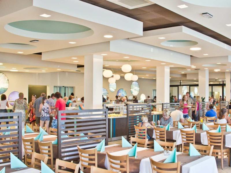 Licht restaurantgedeelte met houten stoelen en gedekte tafels, gasten op de achtergrond.