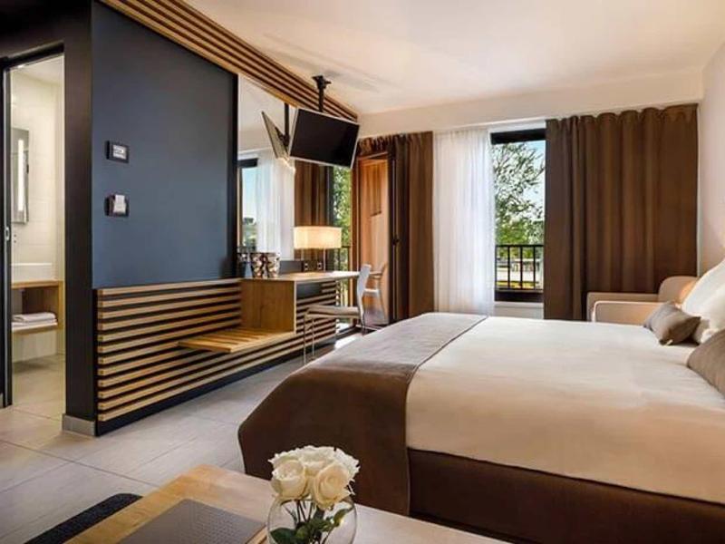 Modern hotelkamer met tweepersoonsbed, tv en uitzicht op balkon met groene omgeving.