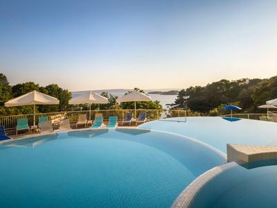 Großer Pool mit blauer Wasserfläche, weißen Sonnenschirmen und Blick auf Pflanzen und Meer im Hintergrund.