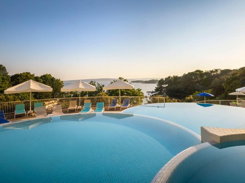 Kruhový venkovní bazén s modrou vodou, obklopený lehátky a slunečníky při západu slunce.