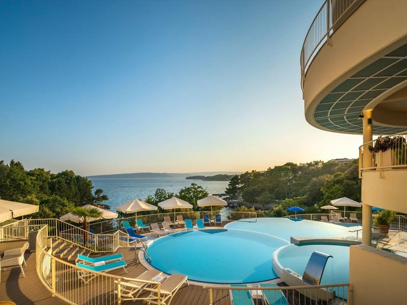 Kruhový hotelový bazén s lehátky a slunečníky, výhled na moře při východu slunce.