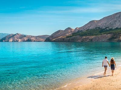 Coppia cammina sulla spiaggia di sabbia dorata vicino al mare turchese con sfondo montano.