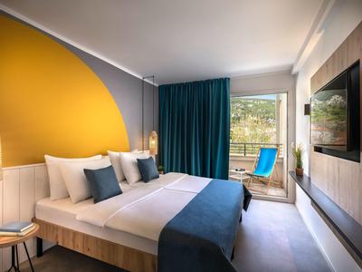 Chambre d'hôtel moderne avec lit double, mur d'accent jaune et accès à un balcon avec sièges.