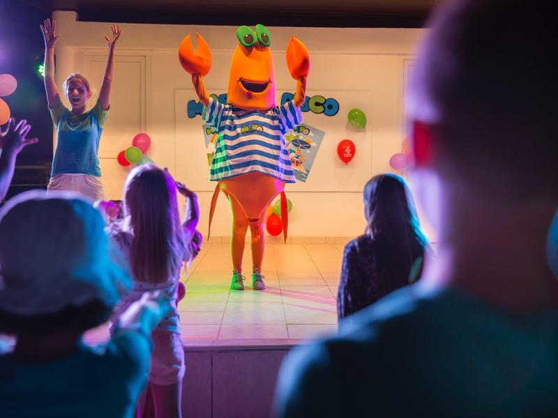 Un groupe d'enfants regarde une performance colorée sur scène avec une mascotte orange.