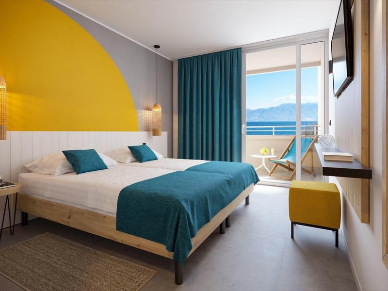 Chambre d'hôtel moderne avec lit double, accents turquoise et vue sur la mer depuis le balcon.