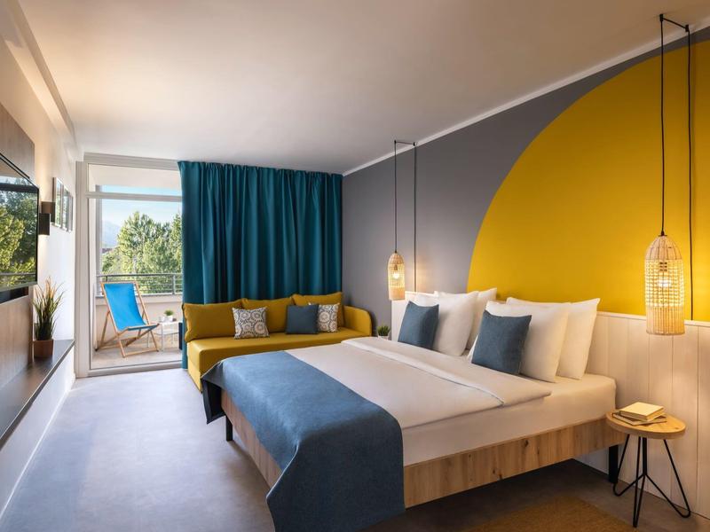 Chambre d'hôtel moderne avec grand lit, décoration murale jaune et terrasse vue sur la nature.