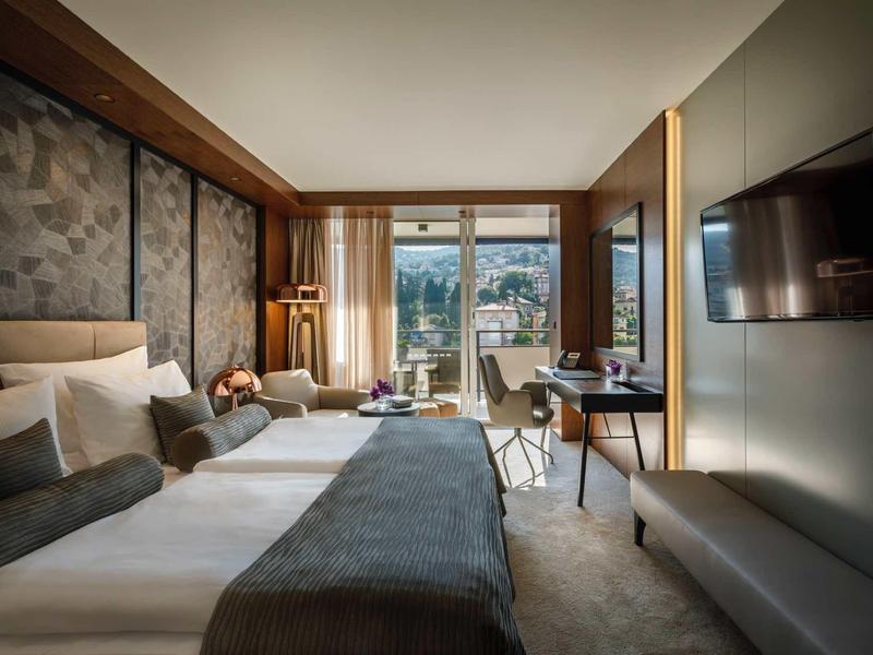 Modern hotel room with large bed, seating area, and city view through large windows.