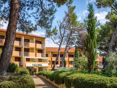 Hotelgebouw met balkons omgeven door bomen en verzorgde struiken onder een heldere hemel.