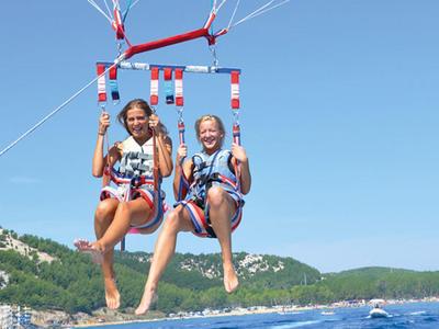 Twee meisjes zitten in een rode parasail harnas boven de zee met heuvels op de achtergrond.