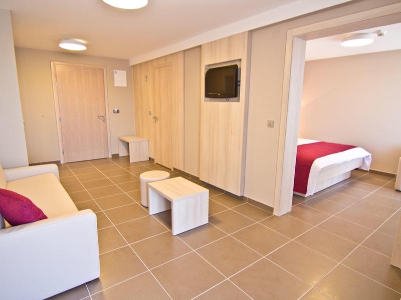 Heller Wohnraum mit weißem Sofa, Stühlen, Fernseher an Wand und angrenzendem Schlafzimmer mit rotem Bett.