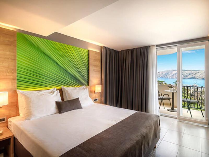 Modernes Schlafzimmer mit großem Bett, grauen Vorhängen, grünem Wandbild und Balkon mit Meerblick.