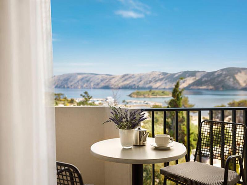 Balkon mit rundem Tisch, Stühlen, Pflanzen, Blick auf See und Berge bei klarem Himmel.