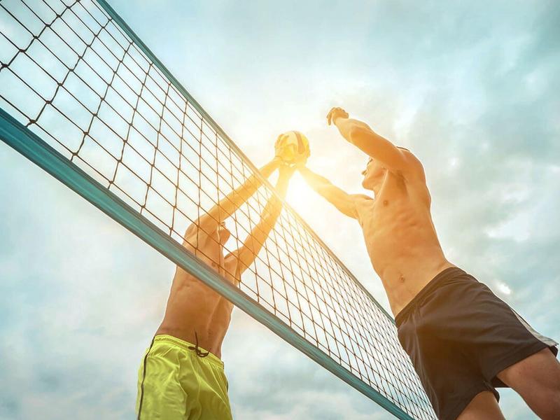 Zwei Männer spielen Volleyball, springen am Netz, Sonne scheint, Himmel bewölkt, sportliche Szene.