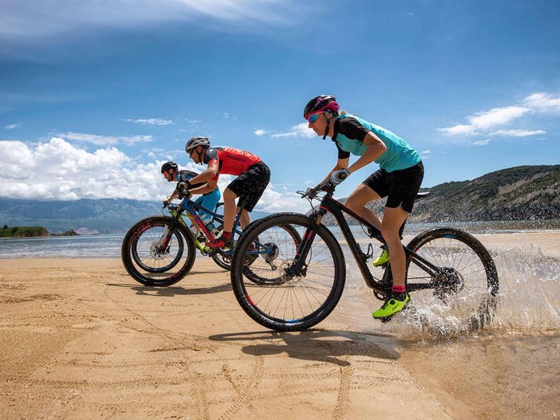 Zwei Radfahrer fahren auf sandigem Strand, blaues Wasser und Himmel im Hintergrund.
