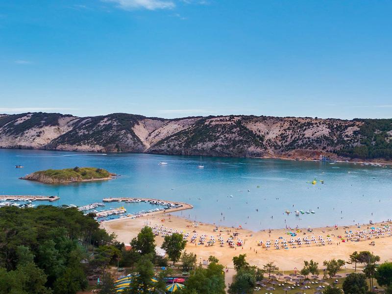 Klares blaues Meer mit Sandstrand, Booten und bewaldeten Hügeln im Hintergrund.