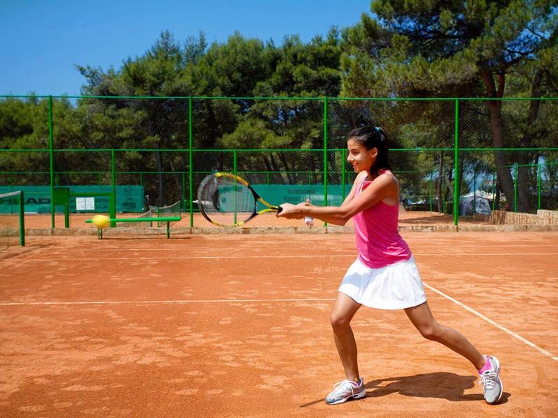 Frau in Sportkleidung spielt Tennis auf rotem Sandplatz bei sonnigem Wetter.